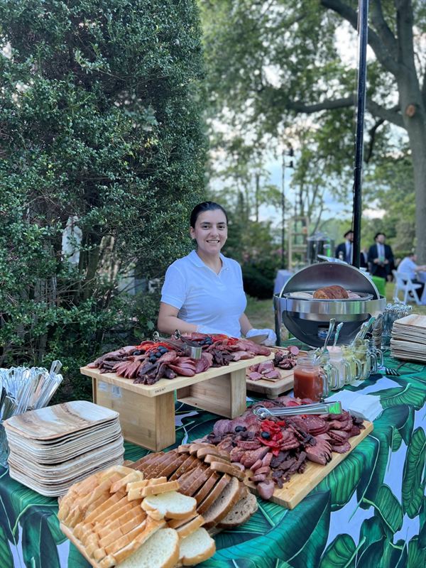 A server in white behind an outdoor deli station: boards of sliced cold cuts, sliced breads, condiments in glass jars and a covered chafing dish.