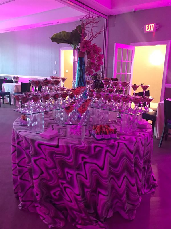 A tiered display table of individual appetizers served in martini glasses, lit magenta.