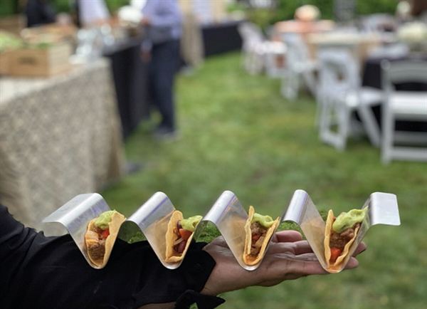 Close-up of five mini tacos in a metal stand, held out on a server's hand at a garden party.