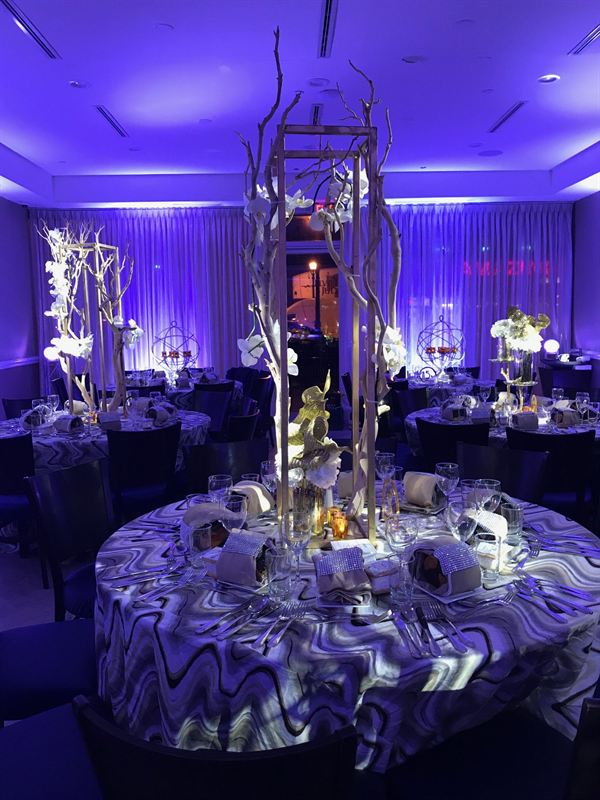 The event room set with round tables, marbled linens and tall gold centerpieces under violet uplighting.